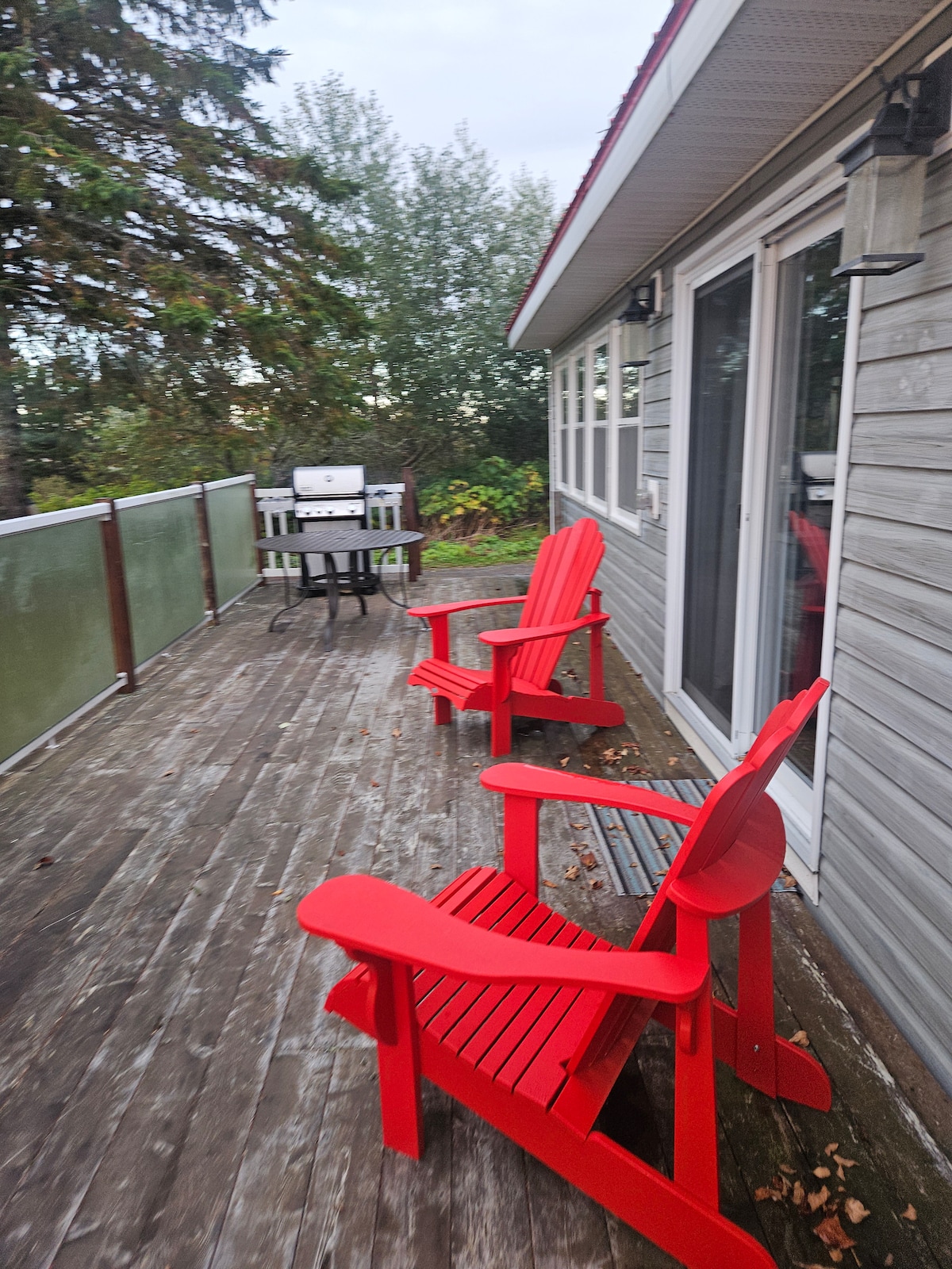 A wooden deck is presented, featuring two red Adirondack chairs positioned for relaxation. A barbecue grill is located near a dining table, with glass panels providing a semi-private atmosphere. Surrounding greenery adds a natural backdrop to the outdoor space.