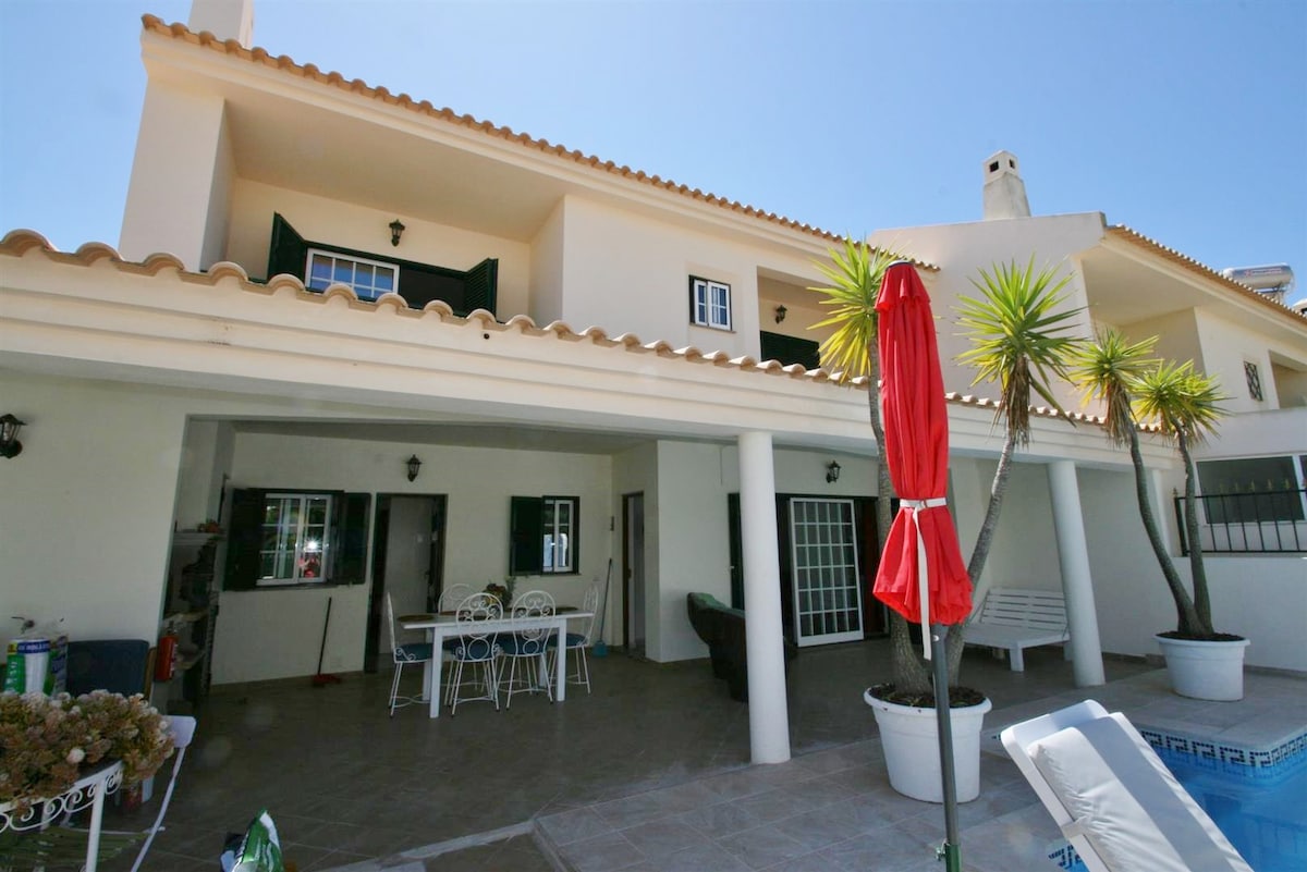 The exterior of the villa showcases a spacious covered terrace, furnished with a dining table and white chairs. Lush palm trees add greenery, while a red umbrella provides shade. The property features a light-colored facade and multiple windows, reflecting a bright and welcoming environment.