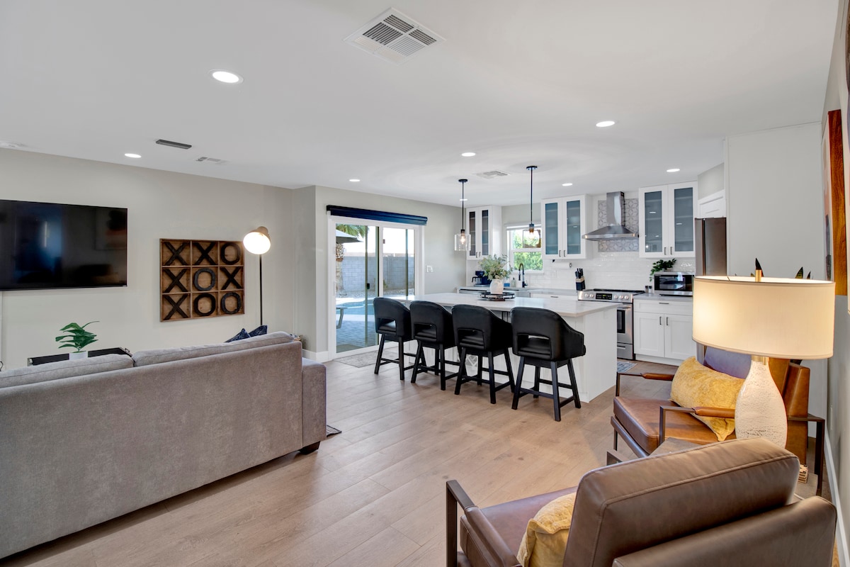 A modern living space is depicted, featuring a comfortable sofa and armchair. A sleek kitchen with bar seating is visible, alongside a dining area. The room is well-lit, with natural light streaming through large sliding doors, which provide a view of the outdoor pool area.