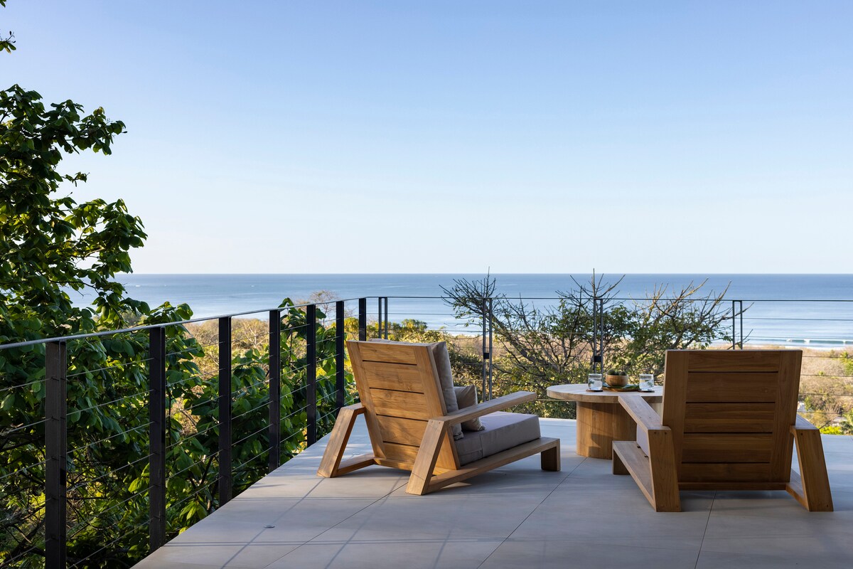 A serene outdoor seating area features two wooden chairs and a round table, overlooking the ocean. Lush greenery frames the view, while the calm blue waters stretch into the horizon under a clear sky.