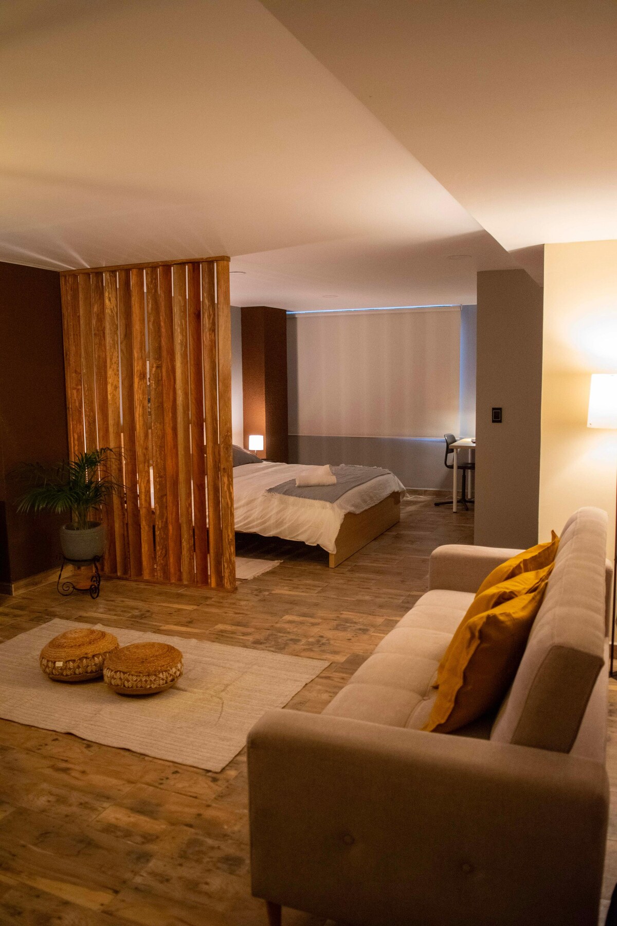 The spacious loft features a cozy sitting area with a neutral-colored sofa facing a light rug. A wooden partition separates the sleeping area, which includes a generously sized bed. Natural light enters through the large window, complemented by soft curtains.