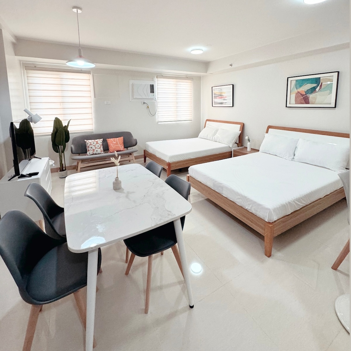 The room features two spacious double beds positioned alongside a modern seating area. A dining table with four chairs is shown, complemented by a sleek television and decorative plants. Natural light filters through two windows with shades, highlighting the minimalist design.