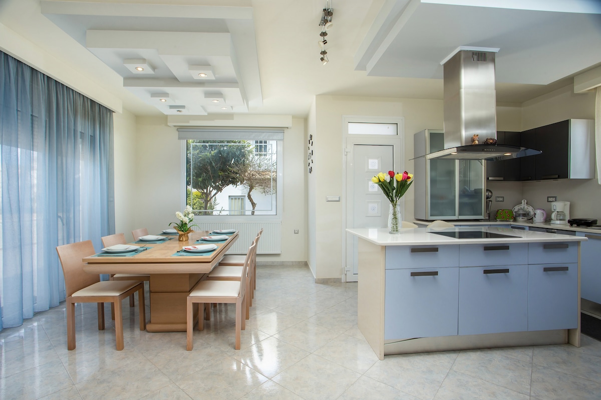 A modern kitchen area is showcased with sleek cabinetry and an island with a cooktop. A dining table with six wooden chairs is placed nearby, complemented by a vase of flowers. Natural light filters through large windows adorned with soft curtains.