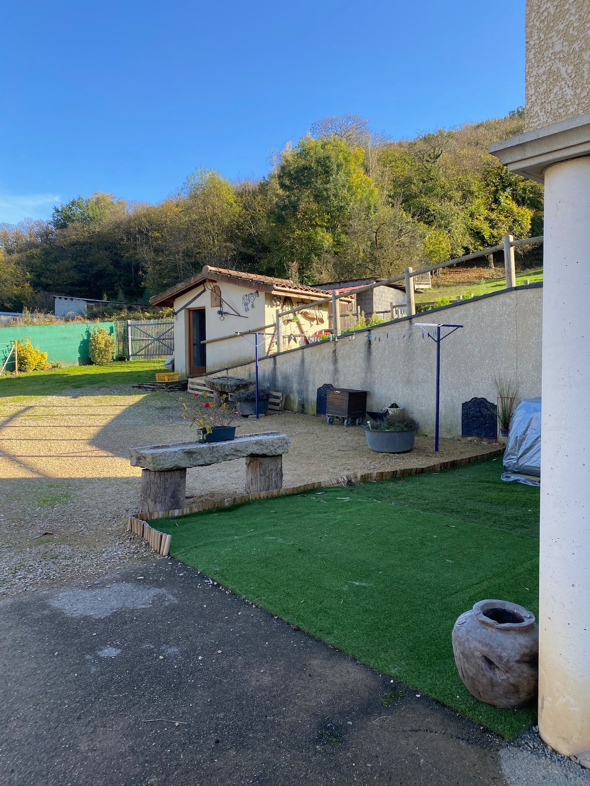 An outdoor area is shown, featuring a gravel path leading to a small building amid greenery. Seating options are available on a patch of artificial grass, with a stone bench and a few decorative items visible. Lush trees and a hillside can be seen in the background.