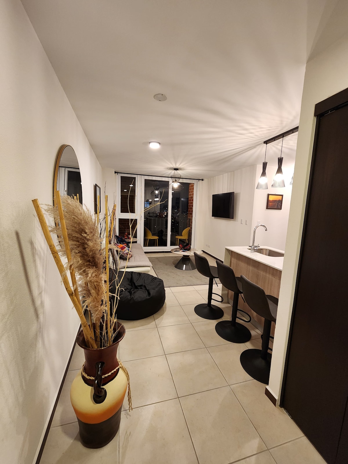 A hallway leads into a modern living space, featuring a circular mirror and decorative plants. High stools are positioned at a kitchen counter, with bar seating visible. Natural light enters through large windows, illuminating the area with a warm glow.