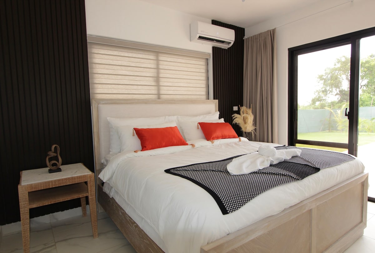 A modern sleeping area features a spacious bed adorned with white linens and contrasting orange accents. Natural light filters through sheer shades, complementing the dark wall paneling. An air conditioning unit is visible above the bed, and outdoor views can be seen through large glass doors.