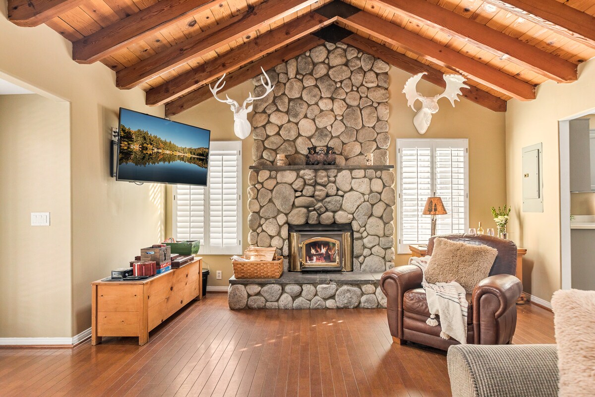 A spacious living room features a stone fireplace as its focal point, with a cozy seating area that includes a leather chair and soft throw blanket. Natural light filters through the window shutters, illuminating the wood-beamed ceiling and warm wooden floors.