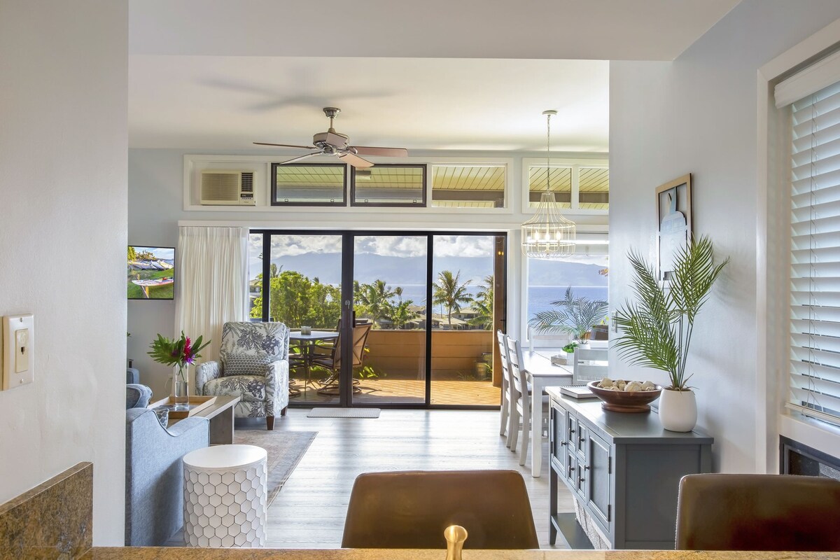 A bright living area features large glass doors that open to a scenic view of palms and mountains. Comfortable seating arrangements are visible alongside a dining area bathed in natural light. The space is enhanced by ceiling fans and contemporary decor, creating an open and airy atmosphere.