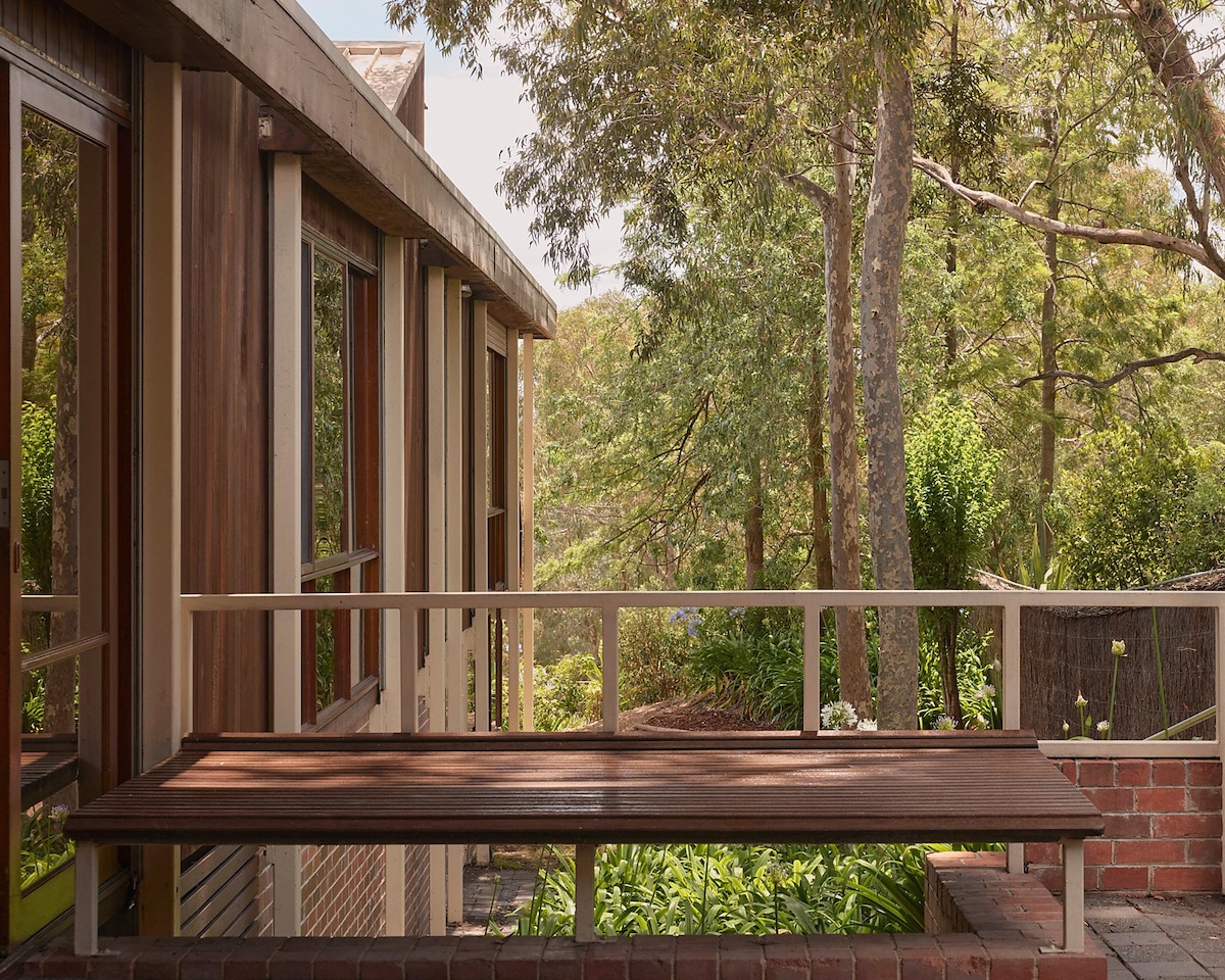 An outdoor balcony is visible at the front of the house, featuring a wooden bench that offers a place to sit and enjoy the surrounding greenery. Tall trees and lush plants create a serene backdrop, enhancing the connection to nature.