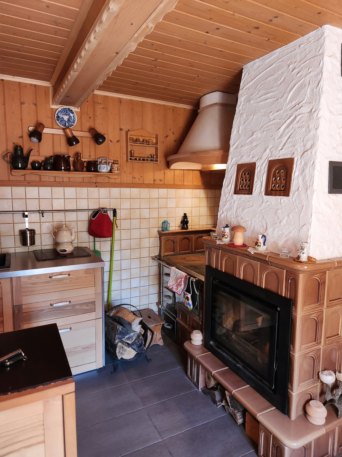 A compact kitchen features wooden cabinetry and tiled walls. An ornate fireplace with a stone facade is positioned beside a vintage-style stove. Decorative pottery and figurines adorn the shelves, adding character to the space. Natural light enhances the warm wood tones throughout.