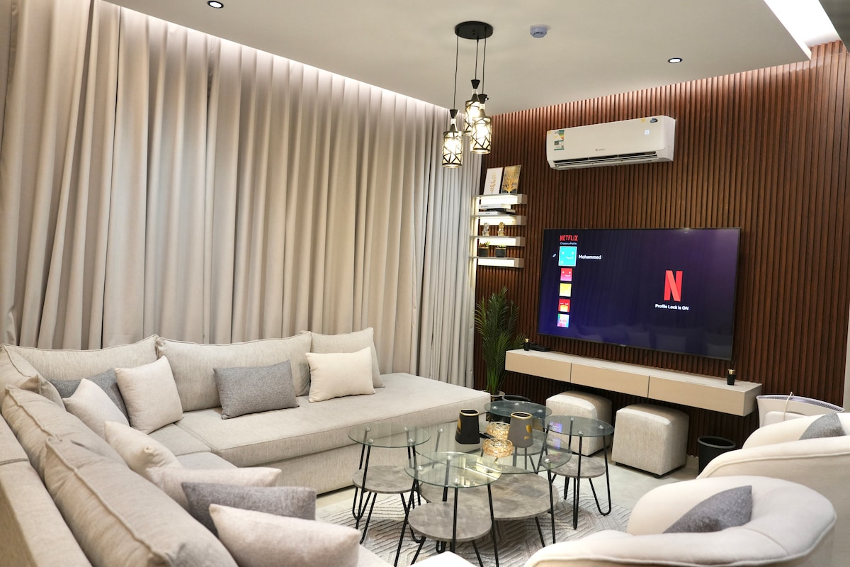 A modern living space is displayed, featuring a large, sectional sofa with light cushions. A glass coffee table is placed centrally, and an 80-inch screen is mounted on a wooden accent wall. Soft lighting from a pendant fixture adds a cozy touch to the area.