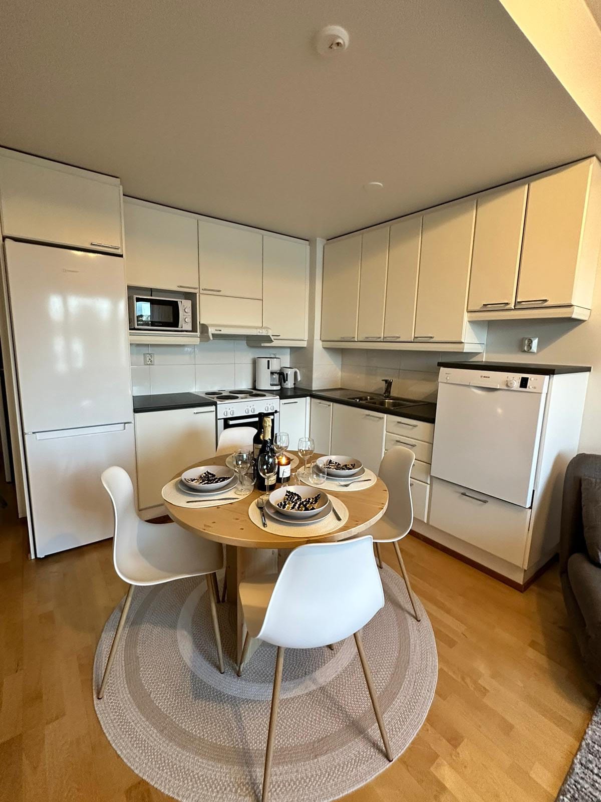 A modern kitchen is equipped with sleek white cabinets and stainless-steel appliances. A round dining table is set for four, featuring plates, glasses, and a bottle of wine. Light wood flooring complements the clean aesthetic, while a cozy rug is situated beneath the table.