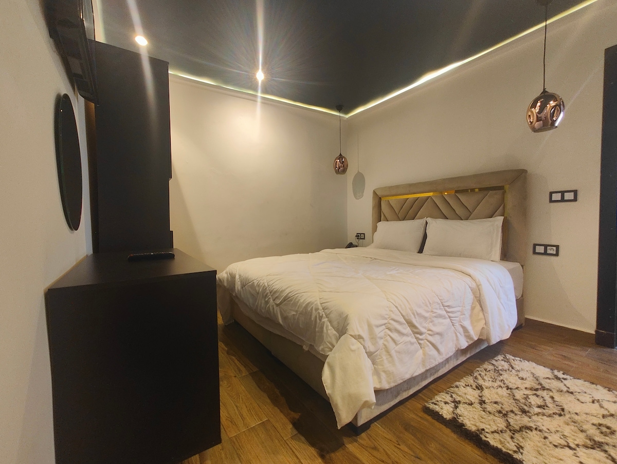 A cozy room features a spacious bed with a plush white comforter. Elegant pendant lights hang from the ceiling, and a sleek storage unit is positioned against the wall. Warm wood flooring and a soft area rug enhance the inviting atmosphere.