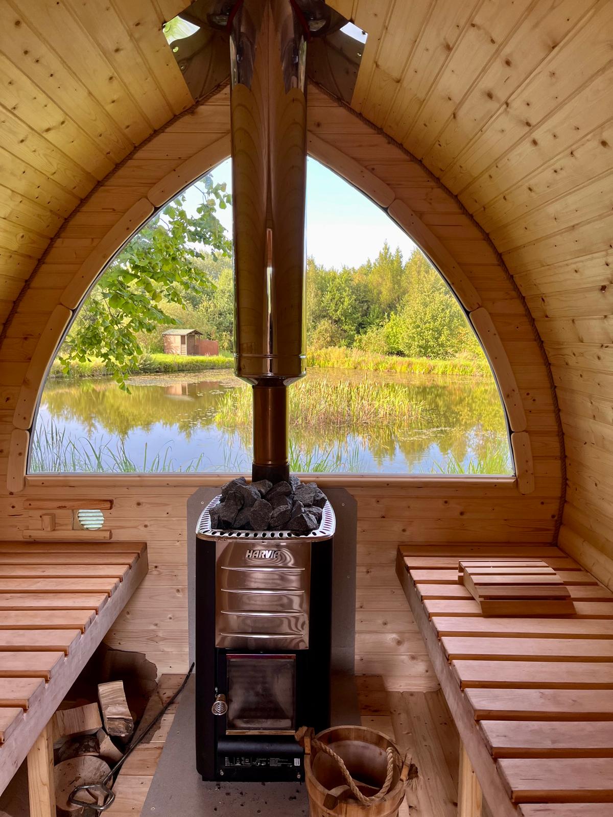 The interior of a wooden sauna features a modern wood-burning stove with a sleek metal chimney. Wooden benches line the walls, providing a space for relaxation. Large windows frame a serene view of a pond and surrounding greenery, promoting a sense of tranquility.