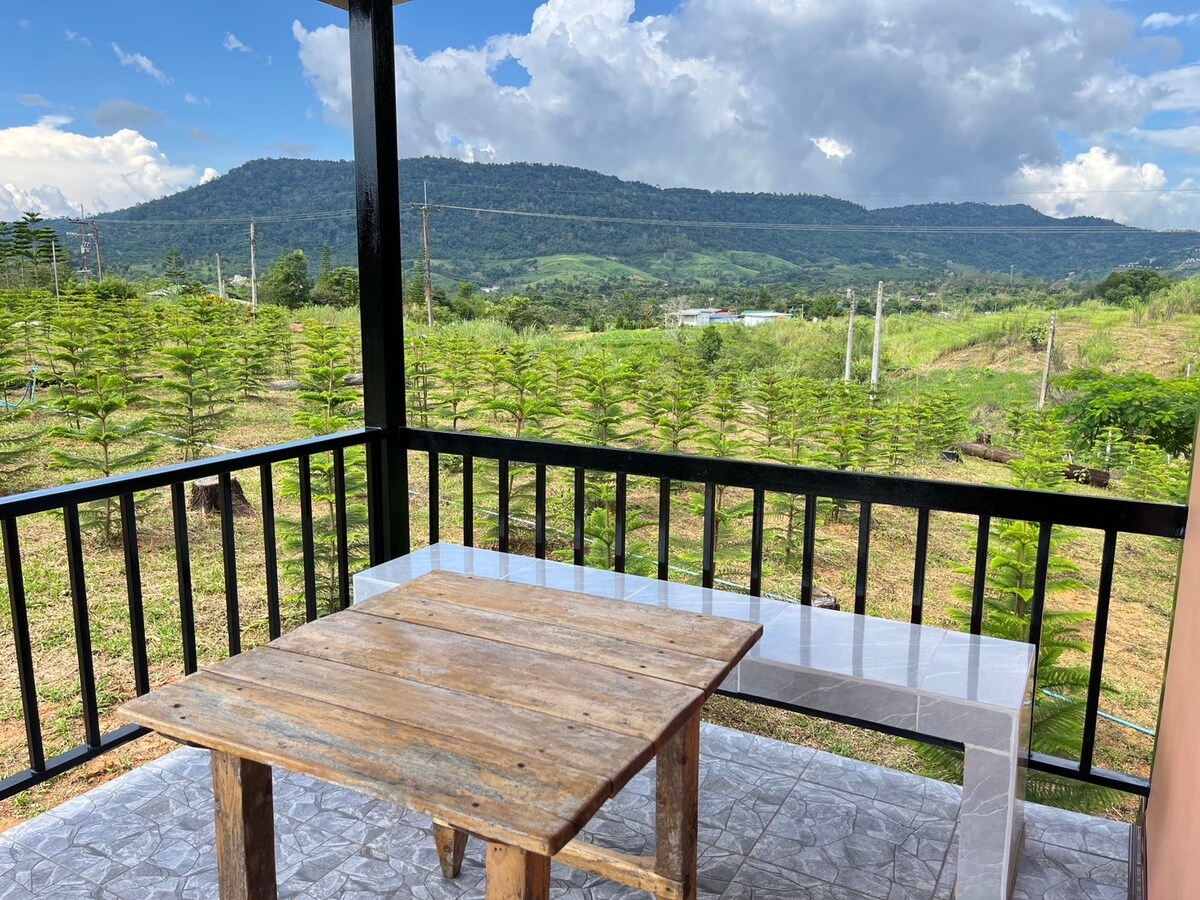 A wooden table is set on a tiled balcony, offering glimpses of lush greenery and rolling hills in the distance. The scenic view includes a mix of trees and open land under a partly cloudy sky, enhancing the tranquil atmosphere of the space.