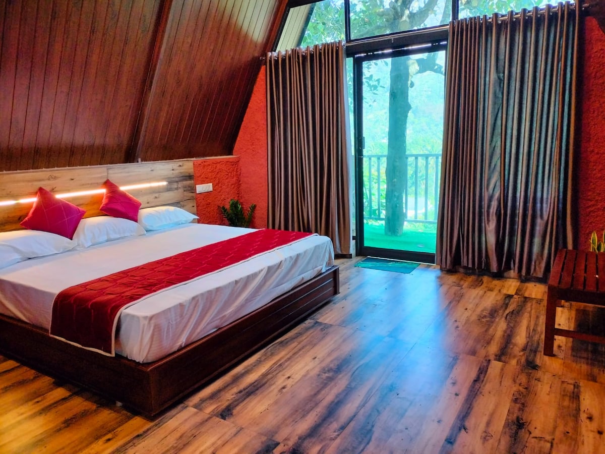 A spacious sleeping area features a large bed with a red and white color scheme and two decorative pillows. Large windows provide natural light and views of the outdoors. The wooden flooring complements the warm tones of the room, while curtains frame the view.