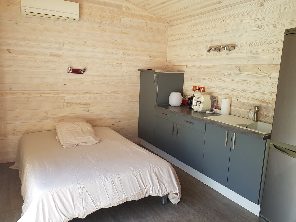 A comfortably furnished studio space features a spacious bed with a neutral-colored cover. A kitchenette is visible, equipped with a stove, sink, and small refrigerator. Light wooden walls provide a natural aesthetic, and a wall-mounted air conditioning unit is installed for climate control.