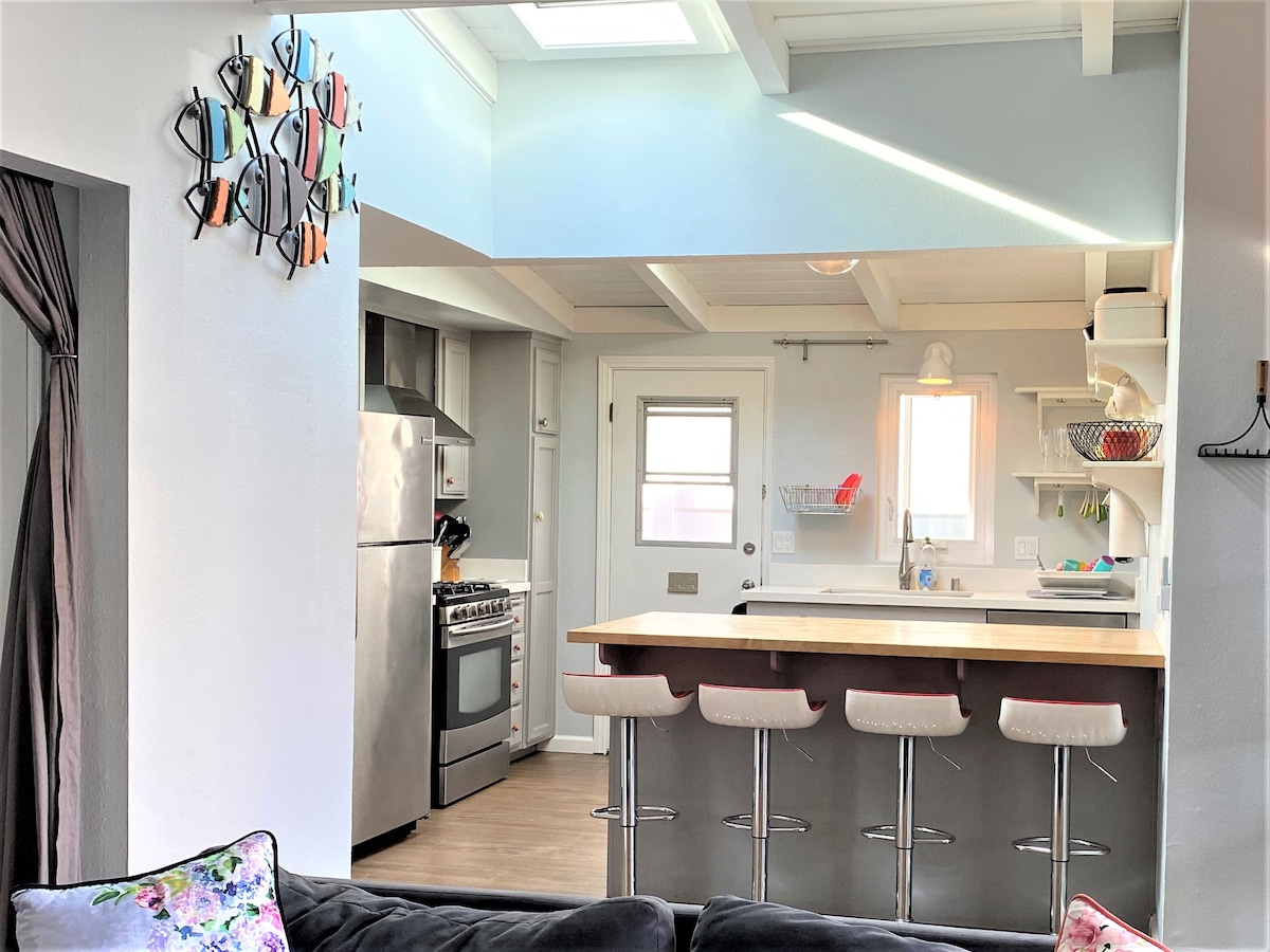 A modern kitchen space is highlighted by stainless steel appliances and a spacious countertop. Three bar stools provide seating at the counter, while natural light is filtered through a ceiling skylight. A decorative wall piece adds a touch of color to the area.