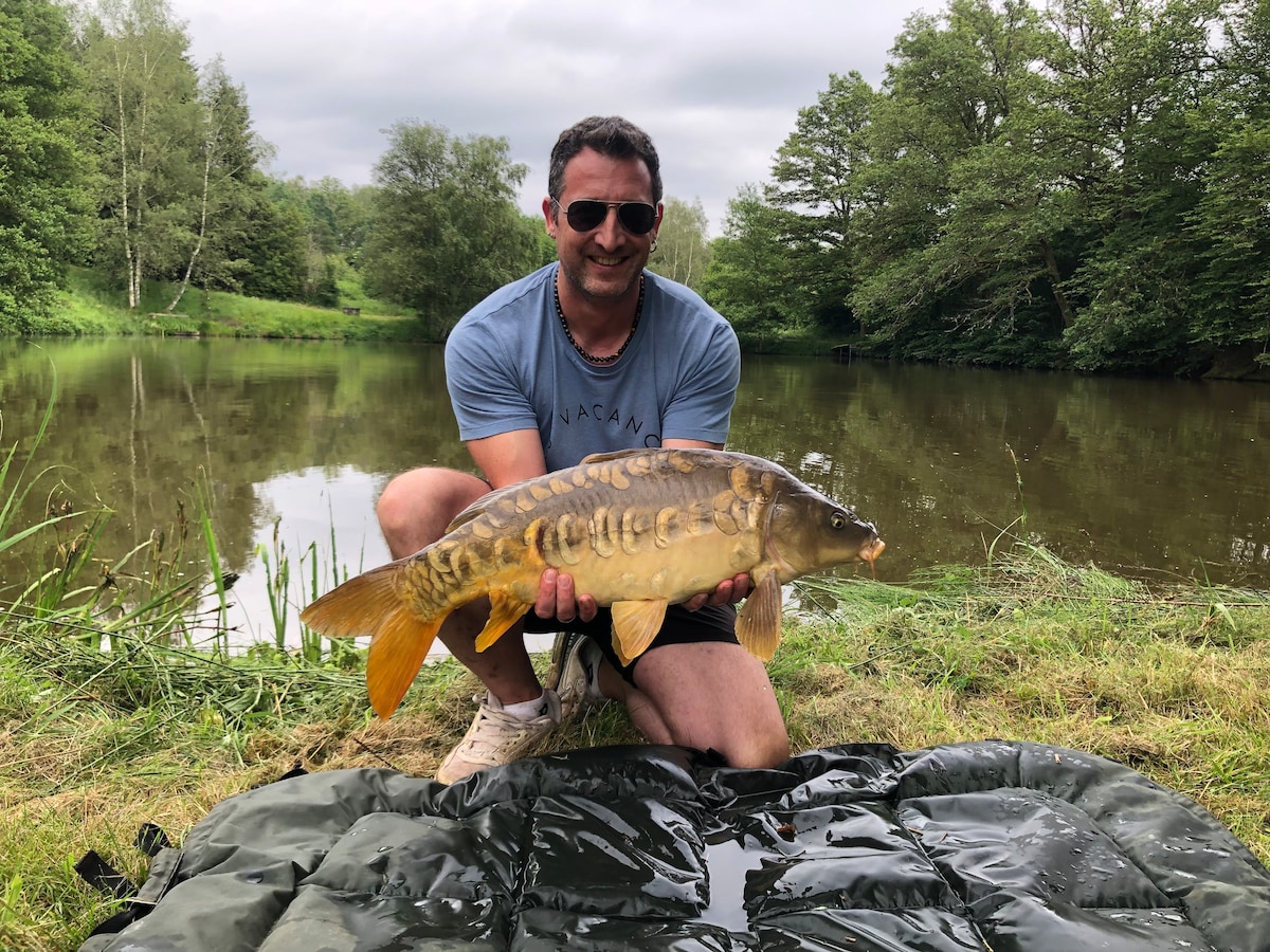 A person is holding a large fish by a serene pond surrounded by lush greenery. The calm water reflects the overcast sky, while tall grasses frame the scene. The fisherman is kneeling on a protective mat, indicating a catch-and-release practice.