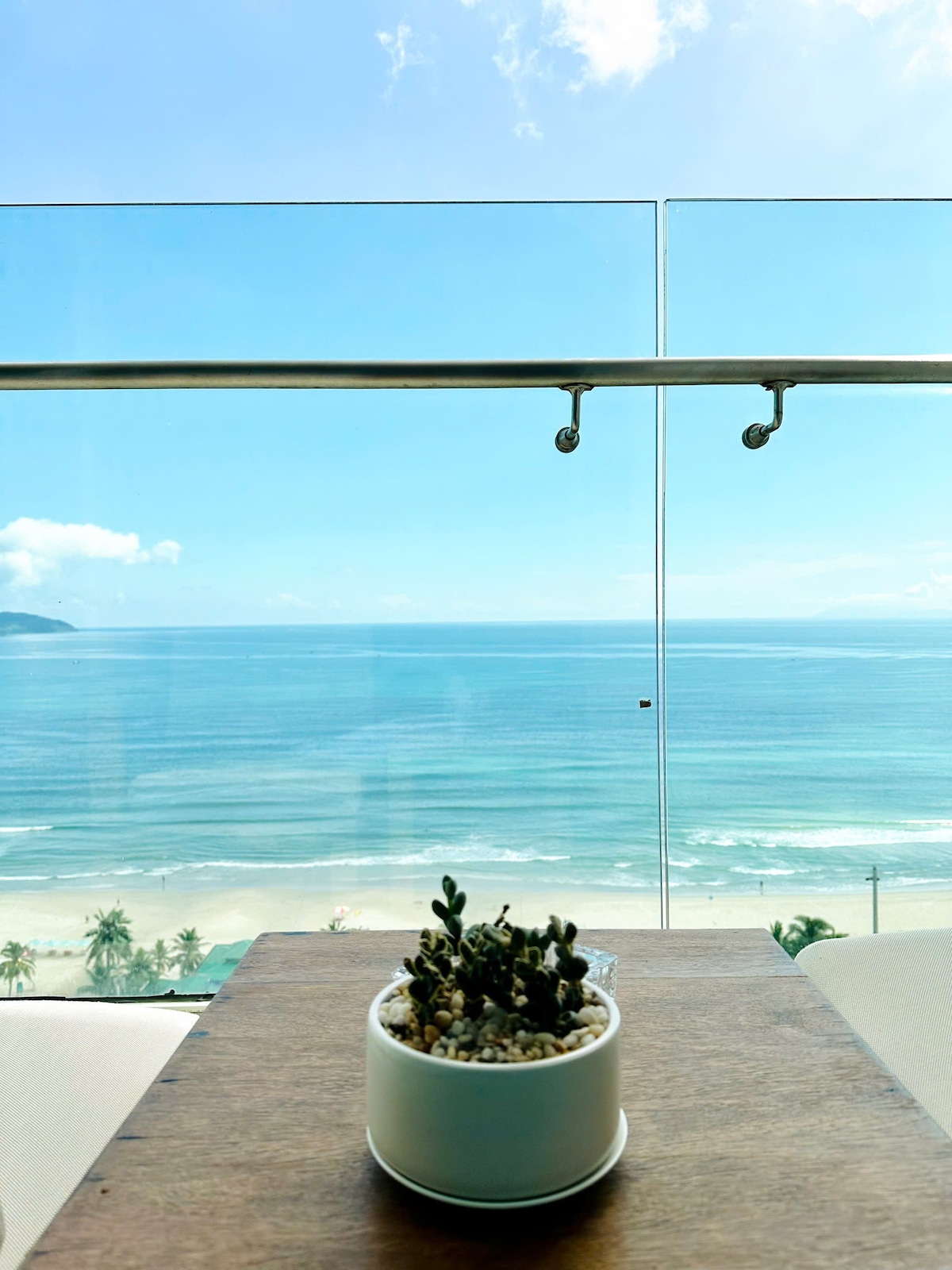 A clear ocean view is framed by a glass railing, showcasing tranquil blue waters and a sandy beach below. A small potted succulent rests on a wooden table in the foreground, adding a touch of greenery to the serene coastal scene.