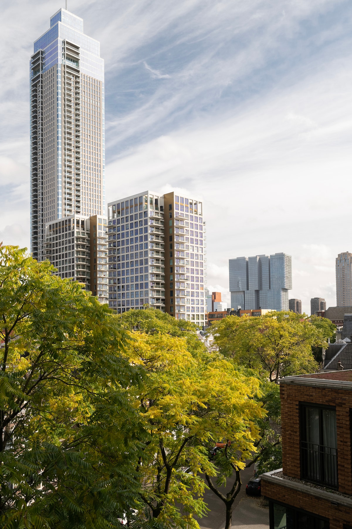 A view from the apartment features a modern skyline with tall buildings, including a prominent high-rise. Lush green trees with golden leaves line the street, contrasting the urban environment. The sky is partly cloudy, creating a dynamic backdrop to the contemporary architecture.