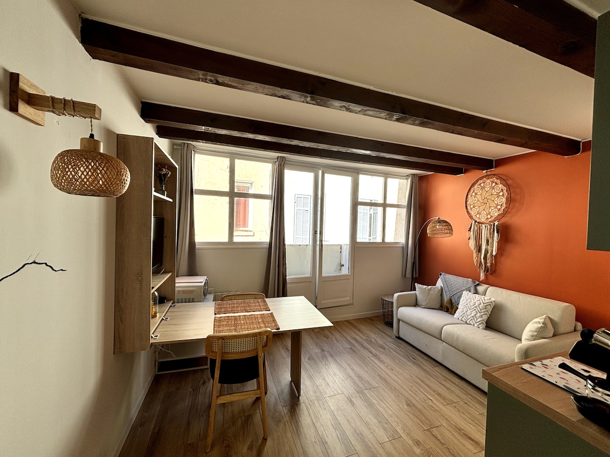 A cozy living space features wooden beams across the ceiling, with a light-toned sofa positioned next to a warm orange accent wall. A compact dining table is visible, accompanied by a chair. Natural light filters through large windows, enhancing the inviting atmosphere.