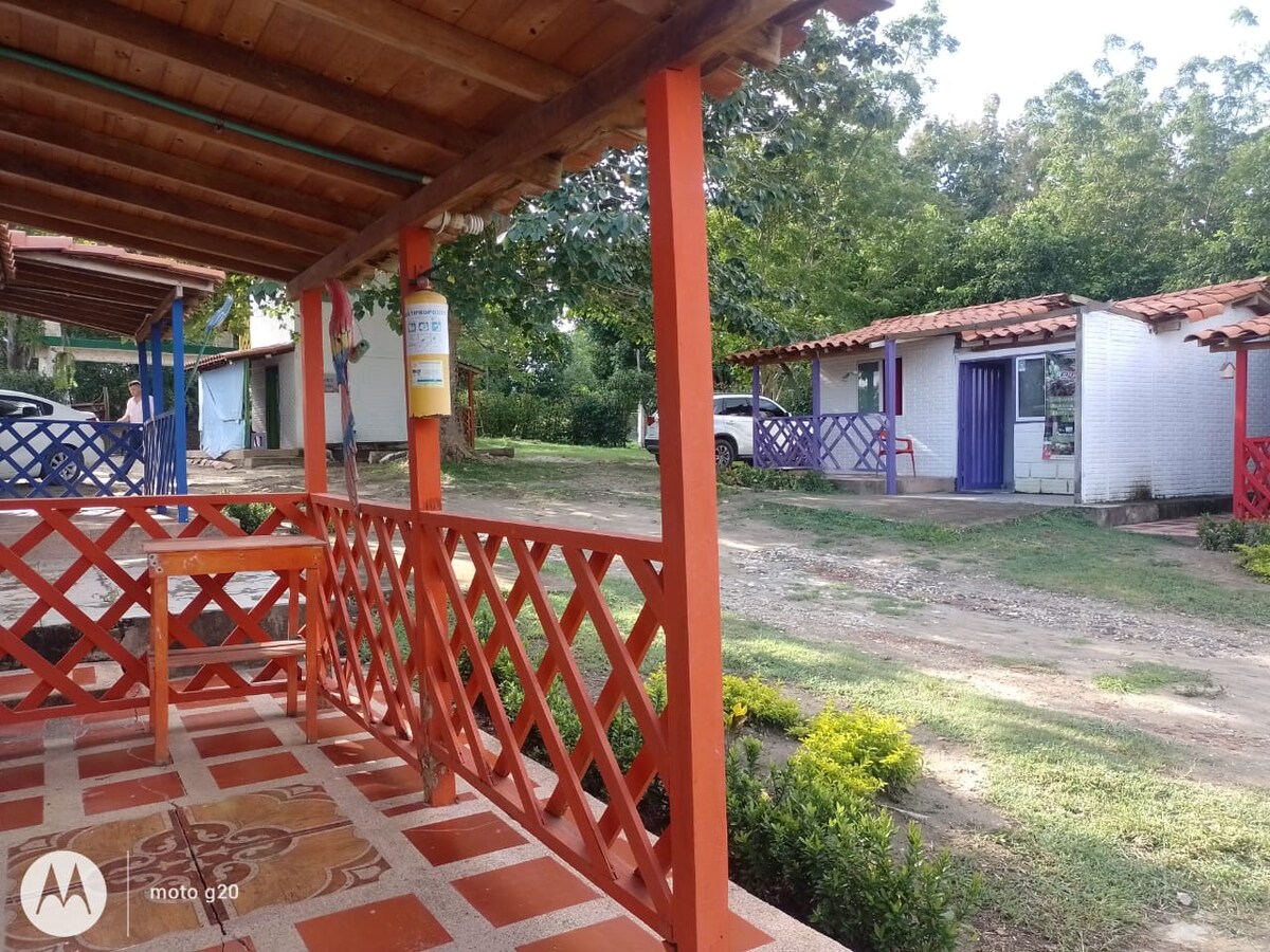Colorful wooden porches, adorned with a crisscross railing design, provide a welcoming entrance to the cabins. The pathways between the cabins are lined with lush greenery, and parking areas are situated nearby, allowing for easy access to the accommodations.