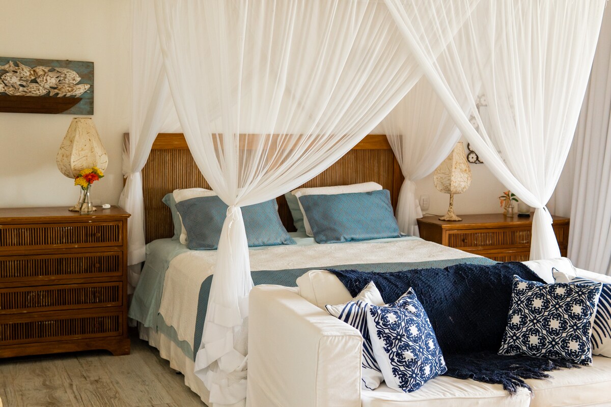 A spacious bed is framed by a delicate draping of white mosquito netting, creating a serene sleeping space. Two nightstands are positioned on either side, each adorned with a lamp. Textured throw pillows and a soft blanket are arranged on a nearby seating area.