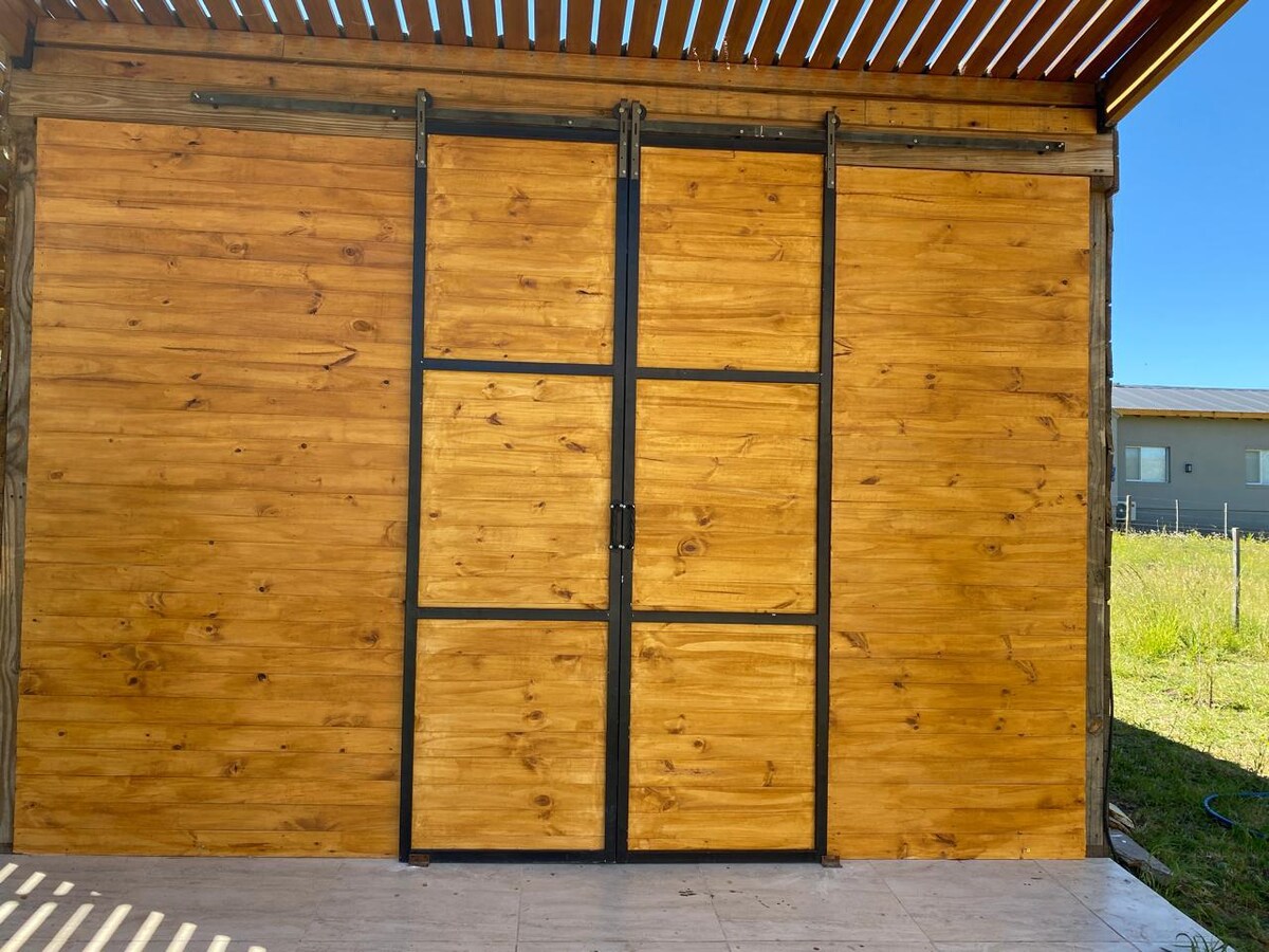 A wooden wall featuring a sliding door is set in a natural environment. The warm hues of the wood contrast with the metal framing of the door. Lush greenery can be seen through the opening, enhancing the connection between indoor and outdoor spaces.
