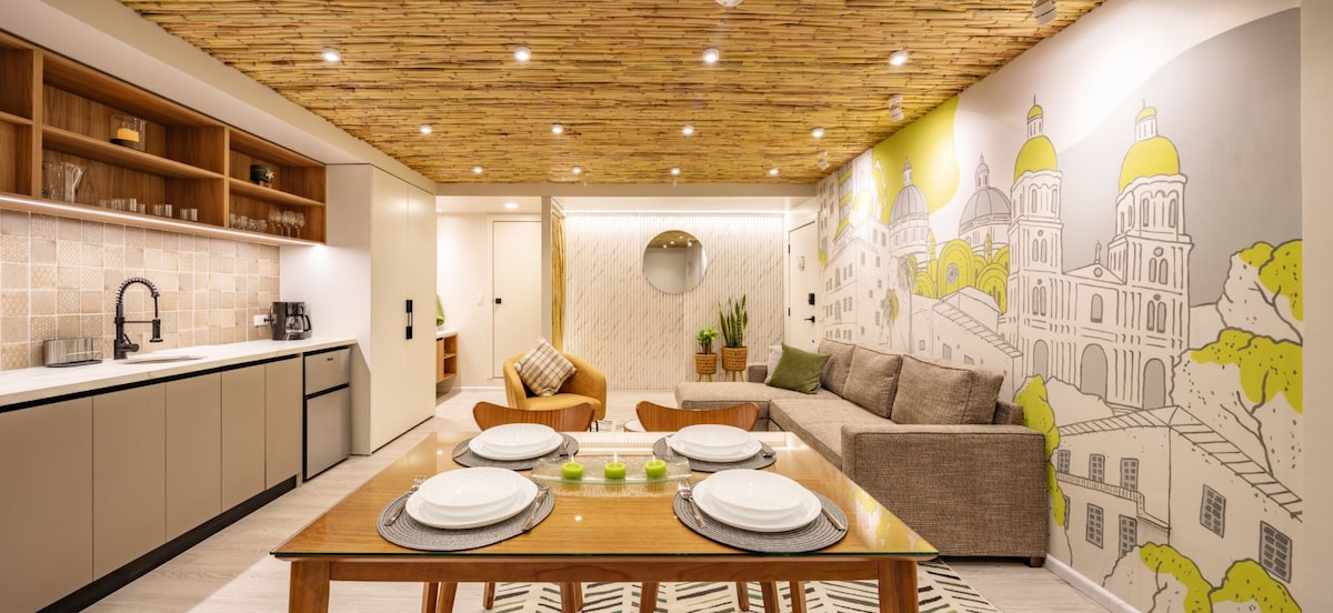A modern living space features a large dining table set with plates and decorative items. A comfortable seating area with a sectional sofa is positioned near a mural depicting architectural elements. Warm wooden textures are present on the ceiling, enhancing the cozy atmosphere.