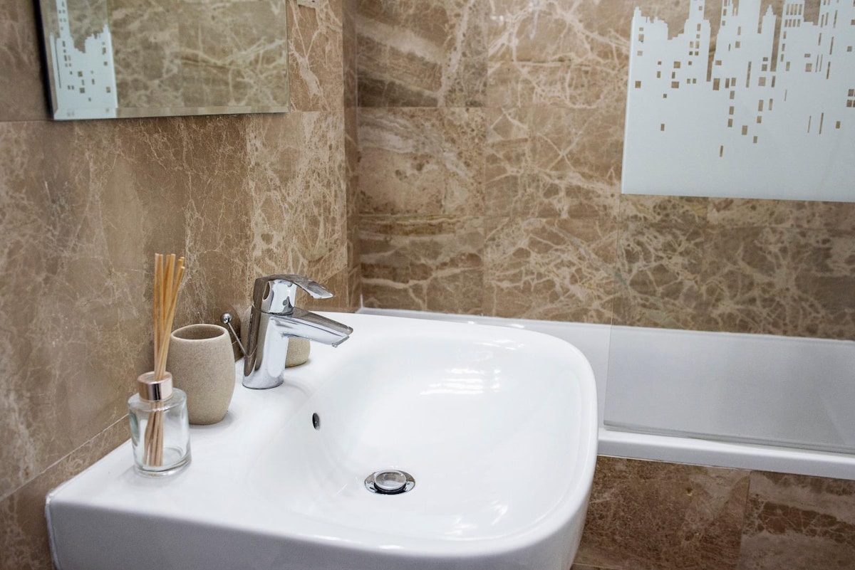 A modern bathroom is presented with elegant marble walls and a sleek design. A white sink is highlighted, accompanied by a stylish faucet. A mirror reflects the space, while decorative items, including a diffuser and a small vessel, are placed nearby.