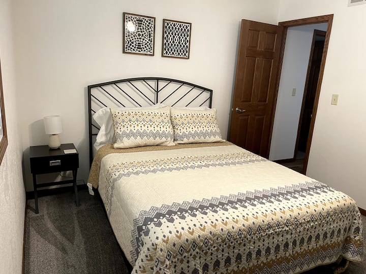 Dormitorio con cama tamaño queen, cómoda, espejo, mesita de noche, lámpara, ventilador de techo, ropa de cama, perchas.