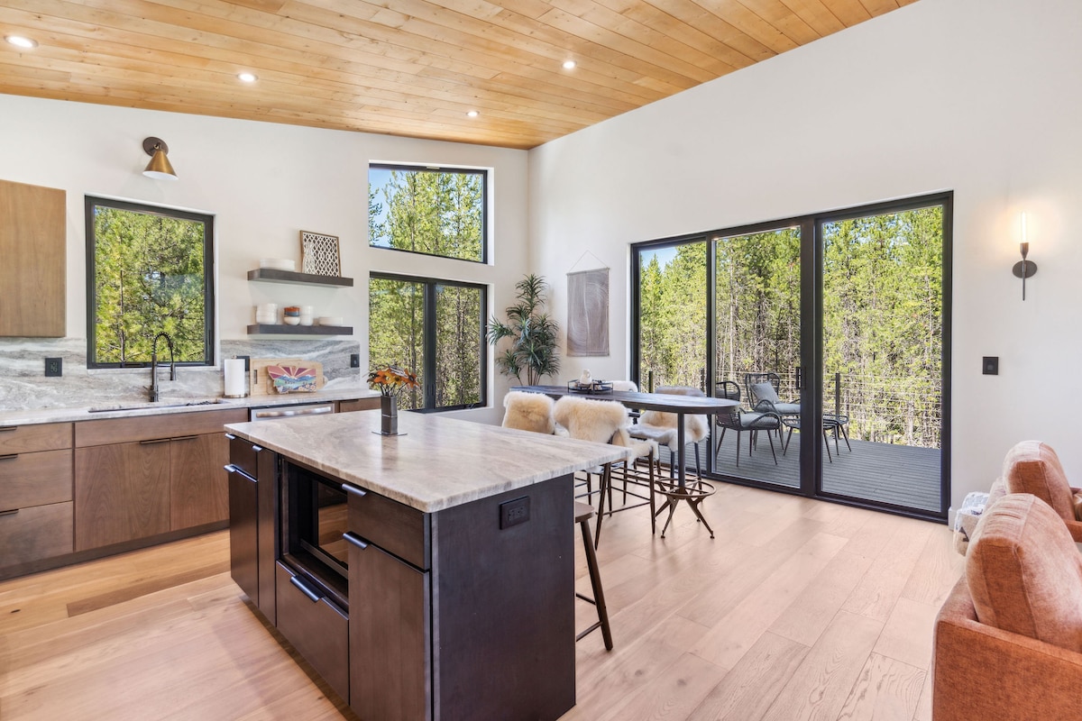 A modern kitchen and living area are presented with natural wood accents and large windows that provide ample natural light. The kitchen island is surrounded by stylish stools, while a comfortable seating area features soft cushions. A scenic deck is visible through the sliding glass doors.