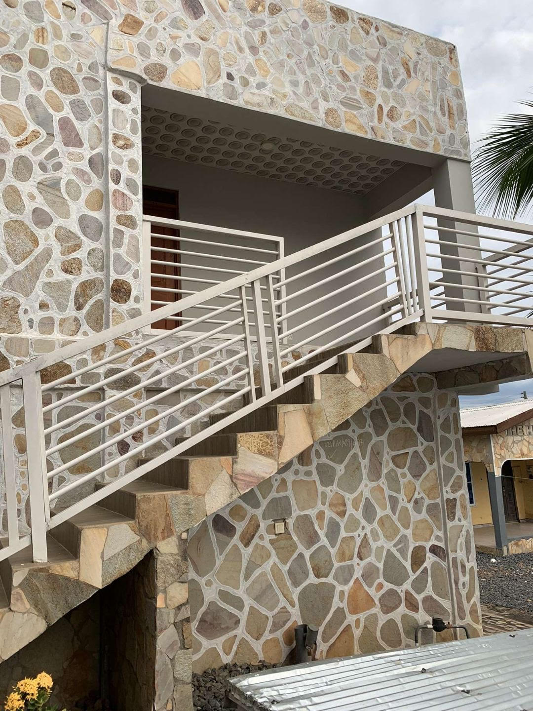 A stone-clad exterior features a set of white metal railings leading up a staircase. The textured surface of the stone contrasts with the simplicity of the railings. Natural light gently illuminates the area, enhancing the inviting appearance of the entrance.