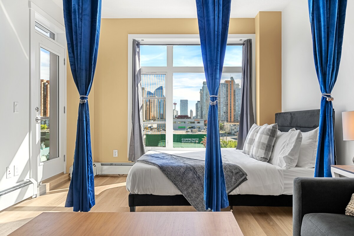 A spacious bedroom features a comfortable bed with a plush gray blanket, framed by large windows dressed in elegant blue curtains. A cozy seating area is visible with a dark sofa. City views, including nearby buildings and the Calgary Tower, are showcased through the windows.
