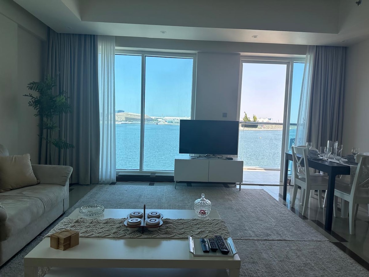 A spacious living area showcases large glass doors that open to reveal a tranquil sea view. A comfortable sofa is positioned beside a modern television stand. A dining table for four is set near the glass doors, allowing natural light to illuminate the room.