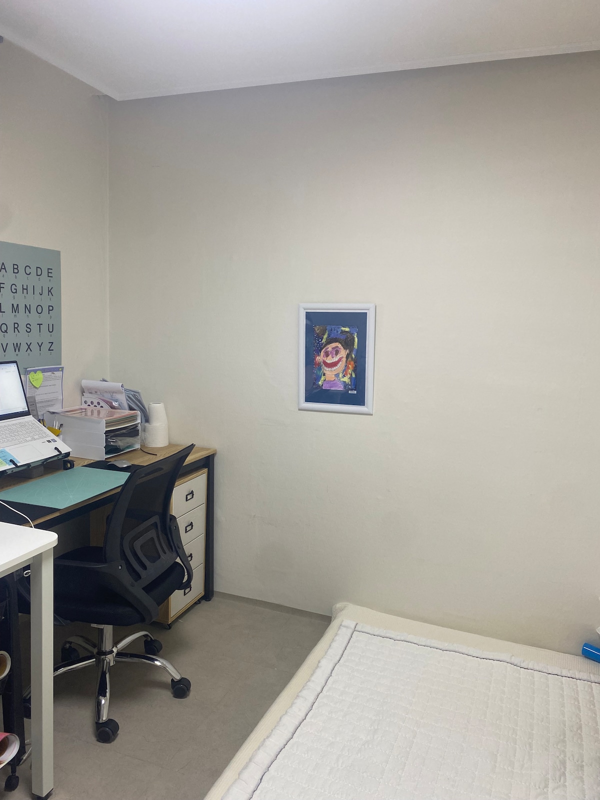 A small workspace is featured with a desk and an ergonomic chair positioned next to a printer and organized documents. A colorful piece of art adorns the wall, complementing the neutral tones of the room. A simple bed is partially visible in the corner.