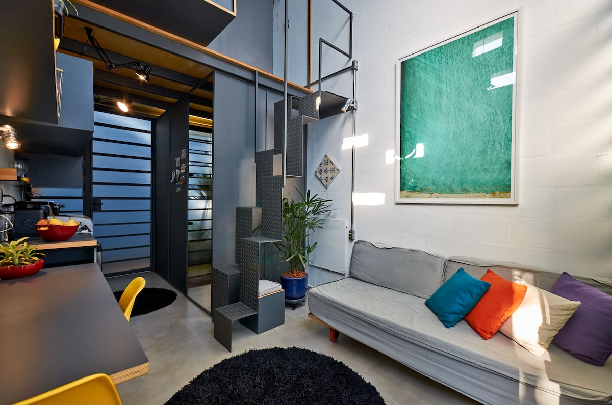 A compact interior showcases a staircase leading to an upper level, with modern, minimalistic design elements. A cozy seating area features a gray sofa adorned with colorful pillows. A wall-mounted artwork in vibrant green adds a pop of color, enhancing the overall aesthetic of the space.