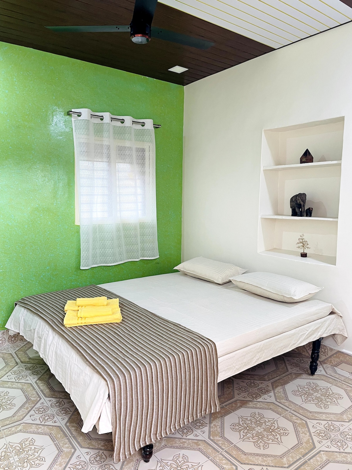 A sleeping area is presented with a spacious bed, adorned with neutral linens and a striped blanket. Soft yellow towels are neatly arranged on the bed. A vibrant green wall adds a pop of color, while light filters through a sheer white curtain hanging above a window.