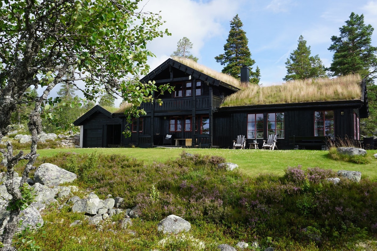 A charming wooden cabin is surrounded by lush greenery and natural stone, featuring a thatched roof and multiple windows. Outdoor seating is provided on the porch, offering views of the landscaped grounds adorned with wildflowers and shrubs.