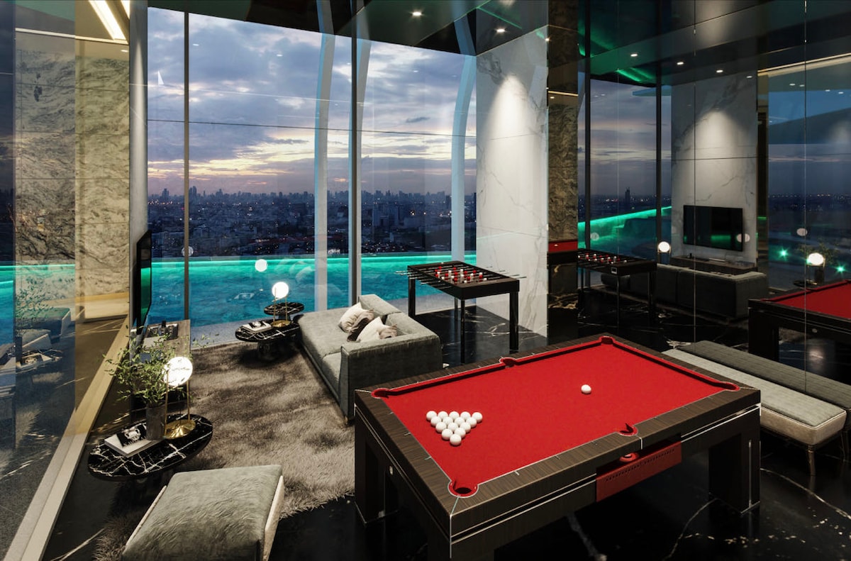 The recreational area features a spacious layout with a billiard table at the center surrounded by comfortable seating. Large windows provide a panoramic view of the city skyline, while soft lighting creates a relaxed ambiance. The décor includes plush rugs and modern furnishings.