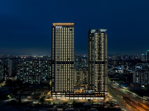The twin towers are illuminated against the night sky, featuring a modern architectural design with numerous windows reflecting city lights. A lively urban surroundings extends beyond, showcasing a blend of residential and commercial spaces in the bustling cityscape.