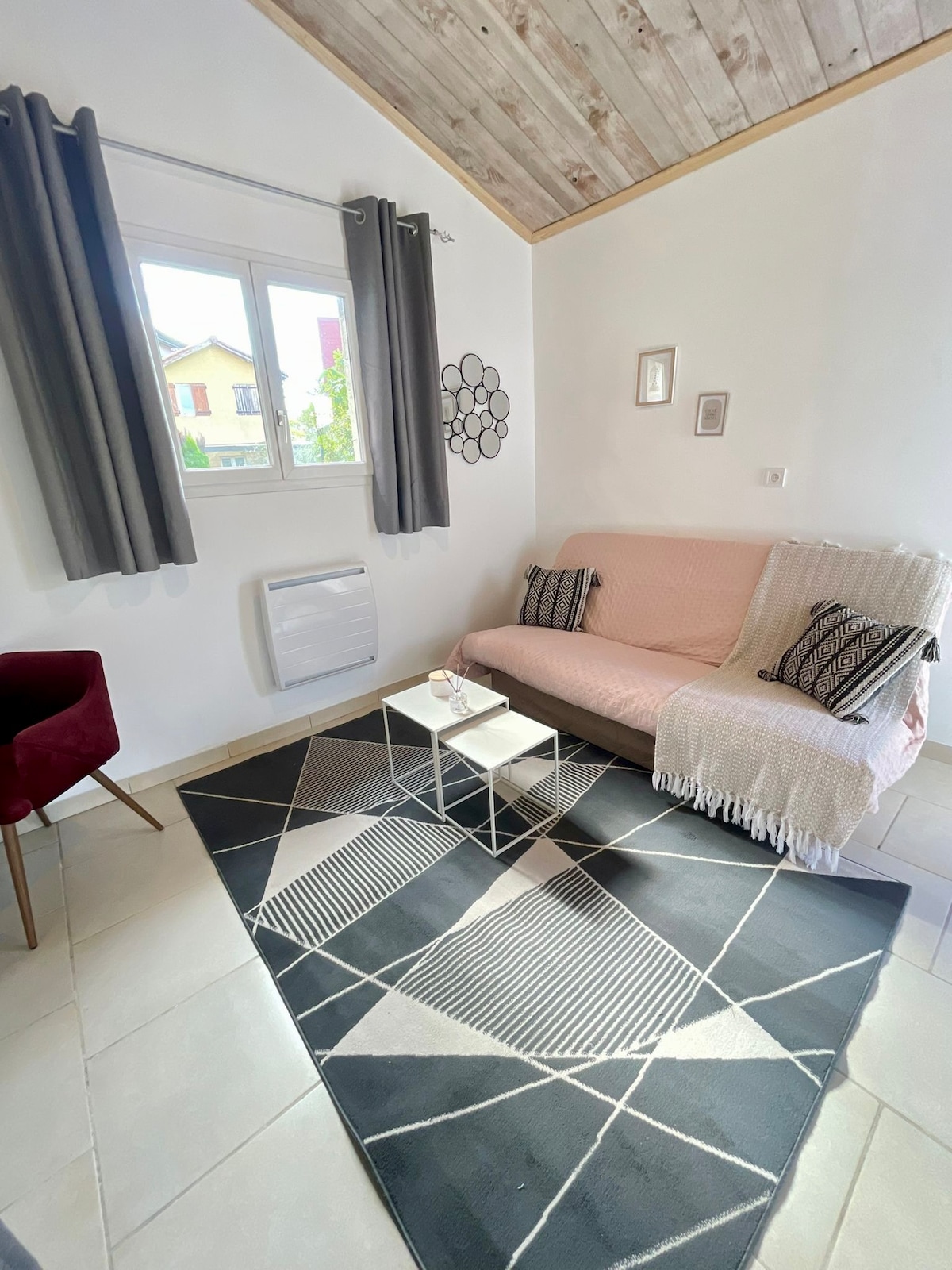A bright living area features a comfortable pink sofa bed and a small coffee table on a geometric-patterned rug. Natural light filters through a window with light curtains, complemented by two decorative wall hangings. A plush armchair in dark red adds contrast to the serene space.