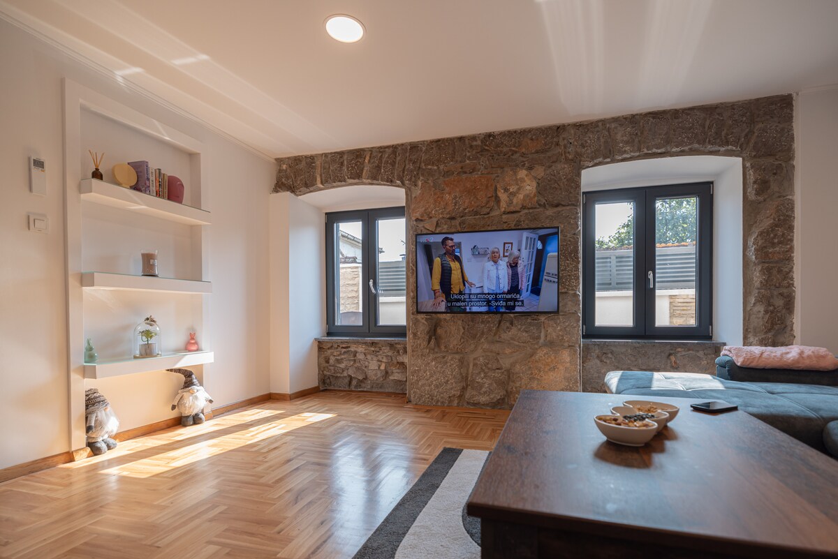 A living area features a large flat-screen television mounted on a stone wall, offering a blend of modern technology and rustic charm. Natural light fills the space through two windows, with a comfortable sofa and a wooden coffee table positioned nearby, accentuated by decorative objects on the shelves.