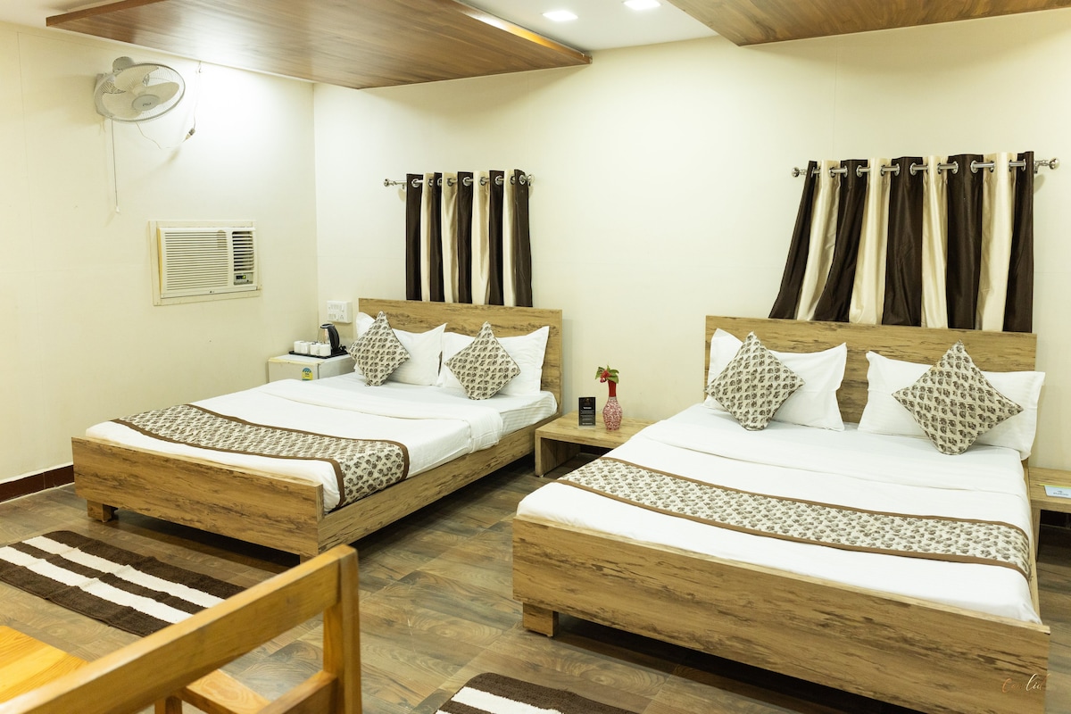 A spacious room with two large beds featuring decorative pillows and crisp linens. Dark curtains frame the windows, while a small fan and an air conditioning unit are mounted on the wall. A wooden table sits nearby on a patterned rug.