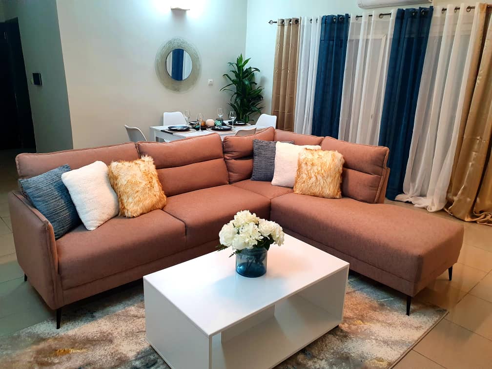A modern sectional sofa in warm tones occupies the living area, accompanied by several decorative pillows. A white coffee table is positioned in front, while a dining table with chairs is visible in the background, softly illuminated by natural light streaming through sheer curtains.