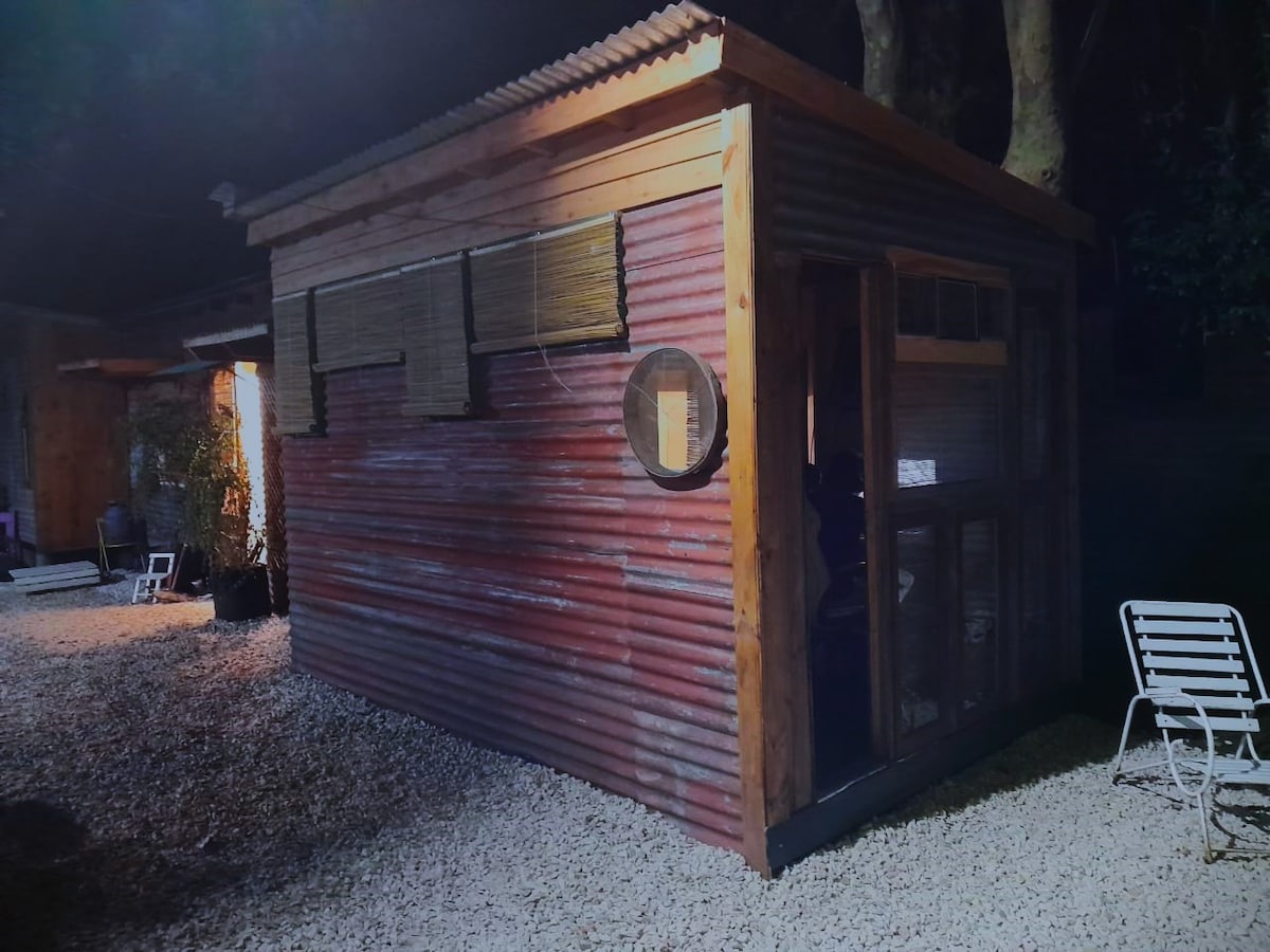 A rustic tiny house is illuminated at night, featuring a corrugated metal exterior with wooden accents. Its large windows provide a glimpse of the interior, while a gravel pathway leads to the entrance. A white lounge chair is positioned nearby.