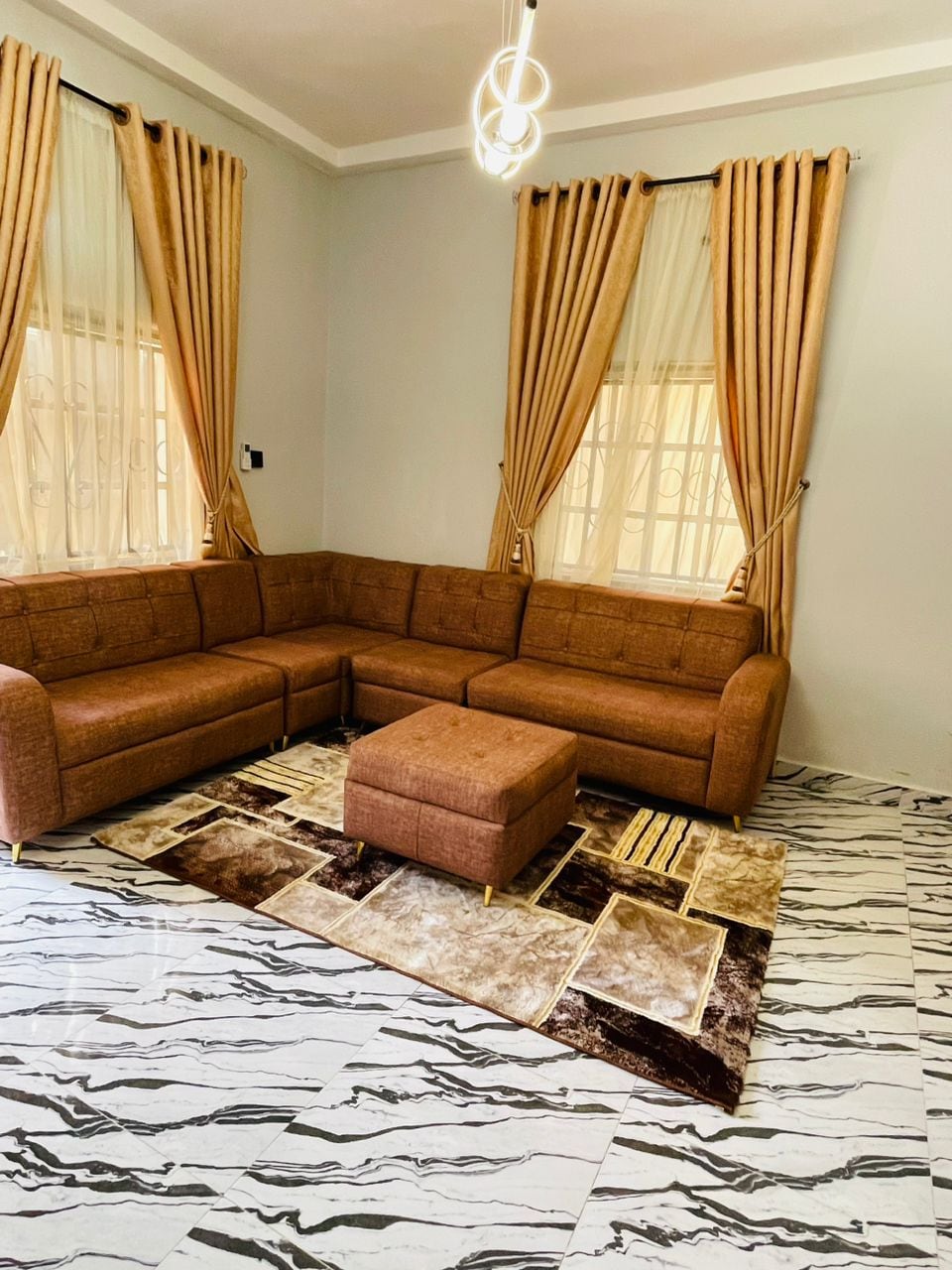 A spacious living area is highlighted by a cozy, brown sectional sofa arranged around a matching ottoman. Natural light filters through sheer curtains on two windows, and a patterned rug adds texture to the marbled floor.