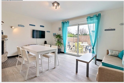 A light-filled living area features a comfortable sofa and a dining table with four chairs. A television is mounted on the wall, while sliding glass doors overlook an outdoor seating area surrounded by greenery. Bright turquoise curtains add a touch of color to the space.