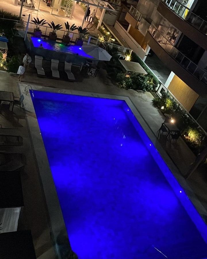 An inviting pool area is illuminated with calming blue lights, surrounded by lush greenery. Lounge chairs are arranged nearby, and a shaded seating area is positioned adjacent to the pool, creating a tranquil outdoor space for relaxation.